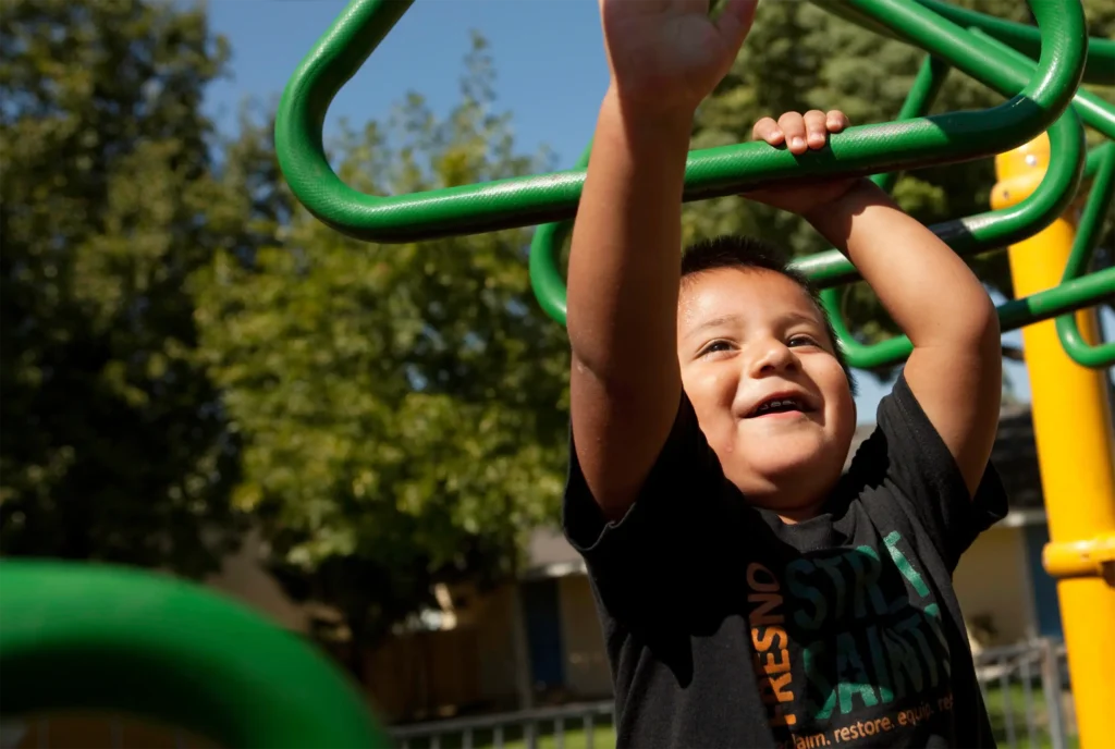 A young child smiles while hanging from green monkey bars at a playground on a sunny day, with trees and a blue sky in the background.