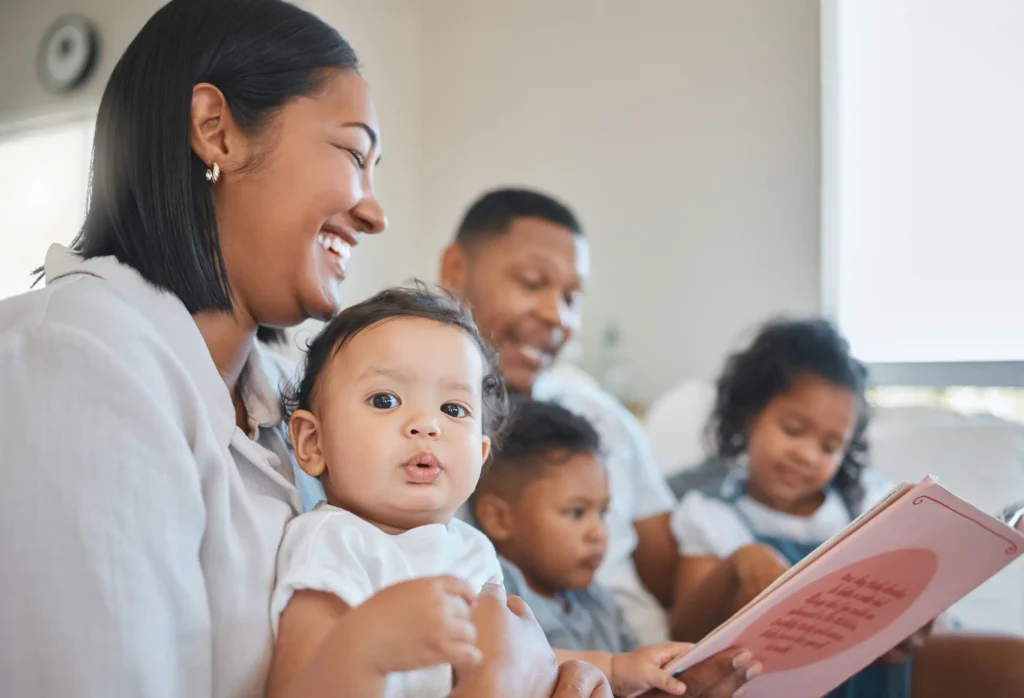 A smiling family of five sits together indoors. The mother holds a baby, while the father reads a book to two young children seated beside him. Everyone looks happy and engaged in the cozy setting.