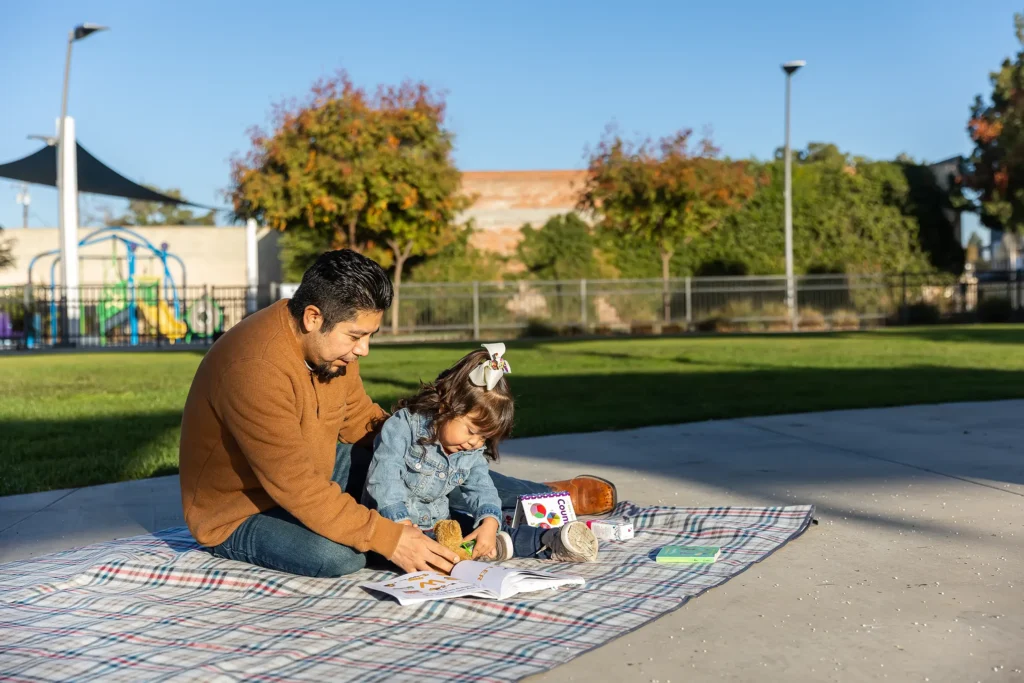 A man and a young girl sit on a blanket in a park, coloring together with art supplies. Playground equipment, trees, and a fence are visible in the background on a sunny day.