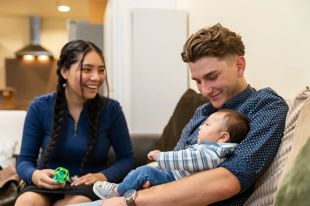 A young man sits on a couch holding a baby in his arms, smiling down at the child. Next to him, a woman with braided hair smiles warmly, holding a green toy ball. The scene is cozy and relaxed indoors.