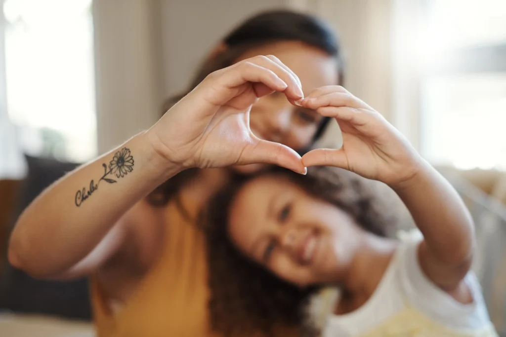 A woman and a child sit close together, smiling and forming a heart shape with their hands. The focus is on their hands, with the woman’s tattoo visible. Their faces are softly blurred in the background.