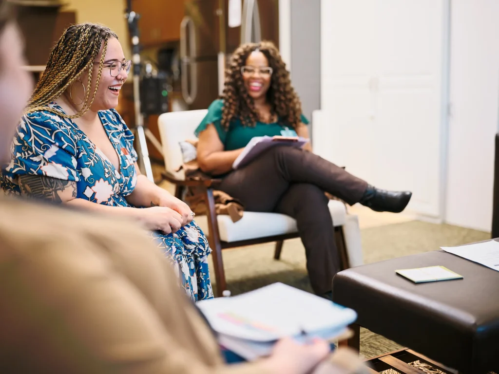 Three women sit in a circle indoors, smiling and talking, with notepads and papers in front of them, creating a warm and collaborative atmosphere.