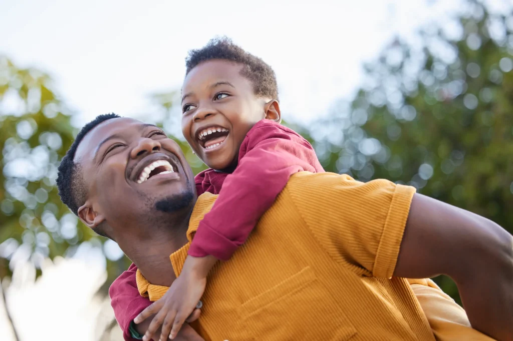 A smiling man gives a joyful young boy a piggyback ride outdoors. Both are laughing, enjoying a sunny day with trees blurred in the background.
